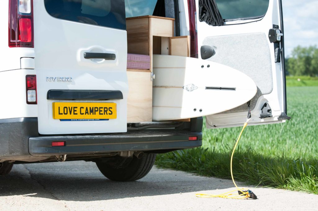 A camper designed for surfing, image features a NV300 Nissan with a surfboard being stowed in the rear.