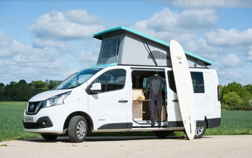 A NV300 with a pop top roof open, the side door open and a surfboard and wet suit on display with the van.